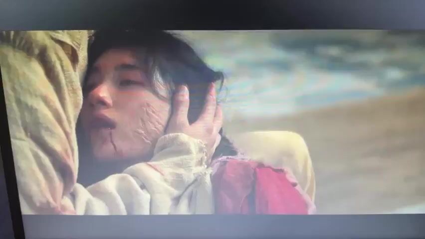 A video shows a woman lying on a light-colored cloth. She has facial scars and blood on her face. A man with long black hair leans over her, his face in distress, and says, "This is your final wish." The setting appears to be outdoors.