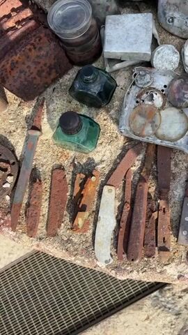 The video shows a close-up, top-down view of various rusty objects laid out on a rough, stone-like surface. The objects appear to be historical artifacts, likely unearthed from a battlefield or historical site.

Dominating the frame are several knives, most of which are heavily rusted, with some showing remnants of their handles. There are also several other metal objects, including what appear to be belt buckles, circular metal pieces, and a few larger, flat metal plates with holes. In the background, there are more metallic objects, some appearing to be containers or parts of larger items. Two small, green glass ink bottles are also visible, one with a black cap and the other uncapped.

The lighting suggests an outdoor setting, possibly during the day, with shadows indicating the sun's position. The ground beneath the stone surface appears to be gravel or dirt. The overall impression is one of a collection of recovered items, possibly from a historical excavation. The audio includes narration in Russian, which mentions a German tank, a village, and a designation of "T3."