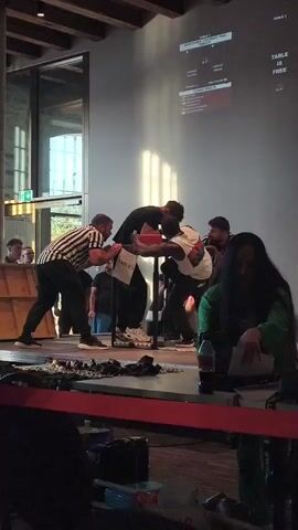 The video shows an arm wrestling match taking place on a raised platform. Two men are actively engaged in the sport, their arms locked on the table, with a referee standing between them, observing the contest. Another referee is positioned on the right side of the table, also monitoring the match. In the background, a large screen displays what appears to be scoring or match information. Several spectators are visible, some standing behind the platform and others seated closer to the front, separated by a red barrier. The environment seems to be an indoor venue, possibly a community hall or event space, with wooden beams visible on the ceiling and large windows in the background. A woman in a green jacket is seated in the foreground, looking down at something in her lap, and a bottle of Coca-Cola is visible next to her. The lighting suggests a daytime event with sunlight streaming in from the left side of the frame.