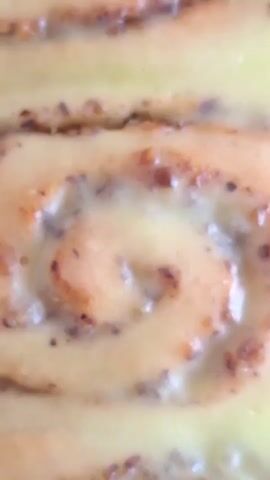 A close-up, top-down shot shows a pan filled with freshly baked cinnamon rolls. The rolls are arranged in two rows of four, showcasing their spiral patterns. A creamy, pale yellow glaze or icing is poured over the warm pastries, filling the spaces between them and pooling slightly on top. The glaze appears to be slightly translucent, allowing the brown, textured filling of the cinnamon rolls to show through. The rolls themselves have a golden-brown hue, indicating they are baked to perfection. Some visible pieces of nuts or spices are embedded in the filling. The overall impression is one of a delicious, homemade dessert ready to be enjoyed.