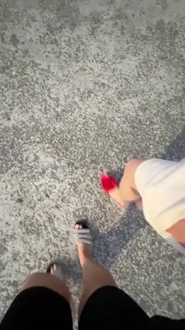 The video shows a first-person perspective of two people walking on a concrete surface. The camera is angled downwards, focusing on their legs and feet. The person in the foreground is wearing black shorts and grey fuzzy slides. The person in the background is wearing a white dress and red fuzzy slides. Their shadows are cast on the ground to their right. The concrete surface appears rough and textured with some small stones visible. The lighting suggests it is daytime, possibly late afternoon or early evening due to the distinct shadows.