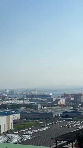 This is an aerial shot of a port with a large cargo ship docked. The ship is white with blue trim and has "EPS PCTC" written on its side, along with a yellow logo. Several rows of white cars are parked on the tarmac below, suggesting a car export facility. Industrial buildings and warehouses are visible throughout the scene, with a hazy blue sky above. The overall impression is one of a busy commercial port during the daytime. There are no people visible in the frame. The location is Pyeongtaek-si, South Korea.