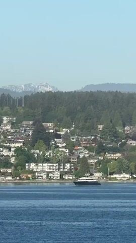 The video shows a cityscape by the water. A large boat is moving across the water in front of the city. The city is densely populated with homes and trees. Behind the city is a dense forest and in the background are mountains. The sky is clear and blue.