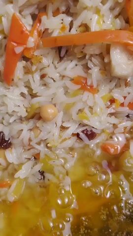 The video shows a close-up of a dish of pilaf being cooked in a large, dark metal pot. The pilaf consists of white rice, shredded carrots, chickpeas, and what appear to be raisins. There are also several pieces of meat, likely lamb, interspersed throughout the dish. The bottom of the pot contains a shimmering yellow liquid, which is likely oil or broth. The camera pans slowly across the surface of the pilaf, revealing the different ingredients and textures. The lighting is somewhat dim, suggesting the dish is being cooked indoors or at night. There are no people visible in the video.