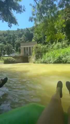 A person is floating in a blue inner tube down a wide river. The river is murky. The person is wearing a life vest and a helmet. There are trees lining the riverbank. Another person in a green inner tube and life vest floats behind. The camera is mounted on the inner tube. In the distance, a building and green, hilly land is visible.