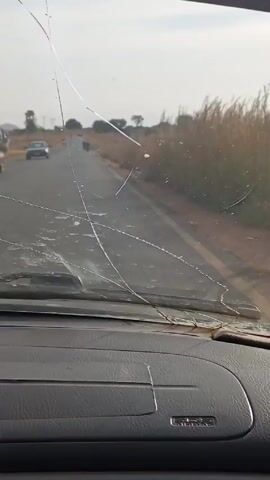 A car is driving down a road. The windshield of the car is cracked. The road is surrounded by fields with some trees and utility poles in the distance. Several motorcycles are driving in the same direction down the road in front of the car.