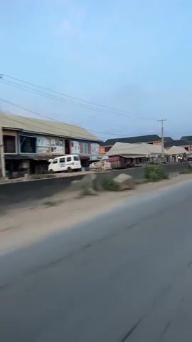 The video shows a view from a moving vehicle on a road. On the left side of the road, there are buildings and a white van. On the right side, there is a highway with a concrete barrier and some vegetation. The sky is visible with some clouds. The scene appears to be in a town or city.