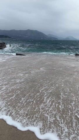 The video shows a concrete platform with waves crashing on it, with a view of mountains in the distance under a cloudy sky. The waves are white and foamy, and they splash against the platform and the rocks at the edge of the water. The sea is a dark, turbulent blue. The platform is gray and damp from the sea spray. The mountains in the distance are hazy.