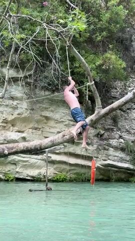 A man is swinging from a rope hanging from a tree branch over a body of water. The man is shirtless and wearing dark shorts. He is holding onto the rope with both hands and swinging forward. The water is a light turquoise color.  Behind the man is a rocky cliff with trees and vegetation growing on top. A life preserver hangs off of the branch of the tree. There are voices cheering the man on.