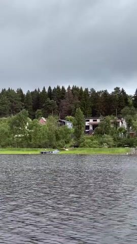 This video shows a view from a body of water, likely a lake or river, looking towards a shoreline. The water is choppy with visible ripples and waves. On the shore, there is a dense line of trees, predominantly evergreens, forming a forest that covers a gently sloping hill. Interspersed within the treeline and closer to the water's edge are several houses, some modern with flat roofs and balconies, others more traditional. There are also areas of lush green grass and reeds along the shoreline. Small docks or piers extend into the water from the shore in a few places, and a couple of small boats are visible near one of these docks. The sky is overcast with gray clouds, suggesting a cloudy or possibly rainy day. The overall scene depicts a residential area nestled within a natural, wooded environment by the water.