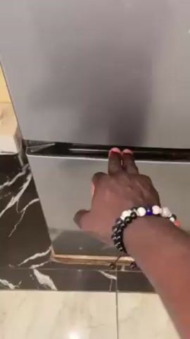 A person with dark skin and a bracelet opens a freezer. Inside the freezer door is a half lemon and a clear plastic container with an orange food product labeled "DORA'S WAJI." The interior of the freezer is otherwise empty. The floor is tiled with a black and white marble pattern.