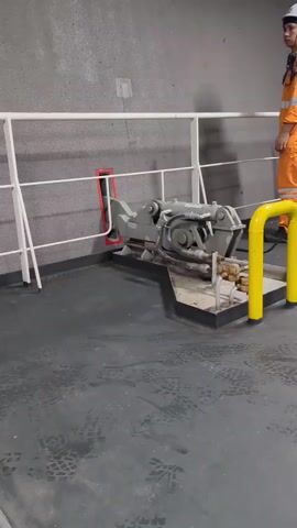 A person in an orange jumpsuit and white hard hat stands to the right of a metal railing. In front of the railing is a complex mechanical device mounted on a gray platform with yellow guardrails. The device is gray and appears to be hydraulic machinery with various hoses and connectors. The background is a rough, gray textured wall. The floor is dark gray with visible footprints. The video is shaky and appears to be filmed from a moving platform.
