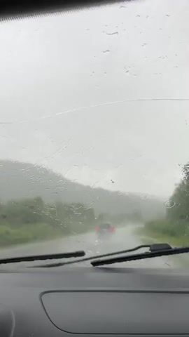 The video shows a view from inside a vehicle looking out through a rain-streaked windshield. Raindrops are visible on the glass, distorting the view. Ahead, a road curves through a landscape of green hills and trees under a cloudy, overcast sky. Several other vehicles are visible on the road, their taillights glowing red in the dim light. The road surface appears wet, suggesting it is raining heavily. The overall mood is somber and somewhat obstructed due to the weather conditions.