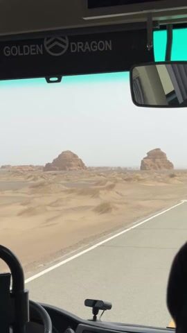 The video captures a view from inside a moving vehicle, likely a bus, driving through a desert landscape in Dunhuang City, China.

**Subjects:** The lower right corner shows a person's shoulder and headrest, suggesting a passenger is filming. A hand is visible on the steering wheel in the lower left, indicating a driver.

**Objects:** Prominently featured are the vehicle's interior elements: a steering wheel, a dashboard, a rearview mirror, and the windshield with a green tint at the top.

**Scene/Setting:** Outside the vehicle, a vast desert stretches under a bright, somewhat hazy sky. The landscape is characterized by sand dunes and distinctive, eroded rock formations known as yardangs. A paved road with a white solid line runs alongside the vehicle.

**Activity:** The vehicle is in motion, steadily driving along the road, offering a panoramic view of the desert and its unique geological features. The camera pans slightly, showing more of the desert.

**Notable Details:** The rock formations vary in shape and size, some resembling large, weathered mounds or ancient structures. The sand dunes show intricate patterns, suggesting wind activity. The overall impression is one of a remote, arid environment. The provided context indicates this is near Dunhuang City, known for its desert landscapes and yardang formations.

**Visible Text:** On the overhead panel above the windshield, the words "GOLDEN DRAGON" are clearly visible next to a logo, likely identifying the bus manufacturer. The audio features indiscernible speech and background noise, consistent with being inside a moving vehicle with people.
