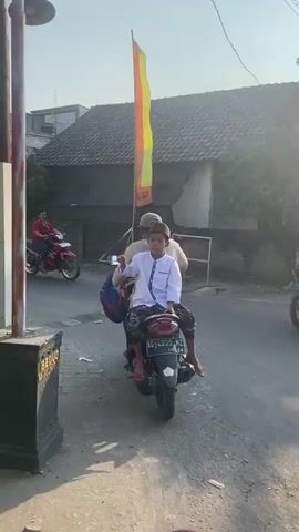 The video shows three people on a scooter. The person in the front is a child wearing a white shirt and a hat. They are holding a long, thin object, possibly a stick or pole, extended in front of them. Behind the child is another person, possibly an adult, also wearing a hat. The third person is out of frame. The scooter is moving down a paved street lined with buildings and trees. There is a tall flag with yellow and red stripes visible to the right of the scooter. The sky is clear and sunny.