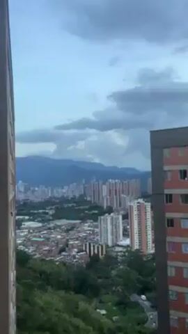 The video shows a cityscape with tall apartment buildings and mountains in the distance. The sky is cloudy and overcast. The camera pans upwards, focusing on a tall brick building and the dark clouds above.