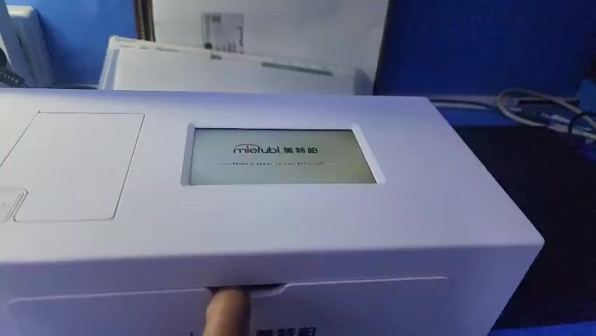 The video shows a white electronic device with a screen. The screen displays text that changes throughout the clip. Initially, the screen is black, then it shows some garbled text, and finally, it displays "mletubol" and some Chinese characters. There is a button labeled "OPEN" on the device. The device appears to be a printer or a similar piece of office equipment. The background is blurry but suggests an office or desk environment with papers and cables visible. The lighting is artificial and gives the scene a cool tone.