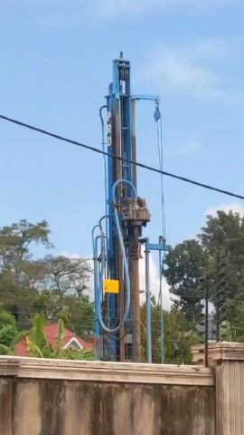 The video shows a blue drilling rig on a property. The rig has the word "PRO" written vertically on it. A yellow warning sign is also attached. The rig is situated behind a concrete wall with an electric fence on top. The background includes trees, vegetation, a red-roofed house, overhead power lines, and a partially cloudy sky. The machine is running, and some birds are flying in the distance.