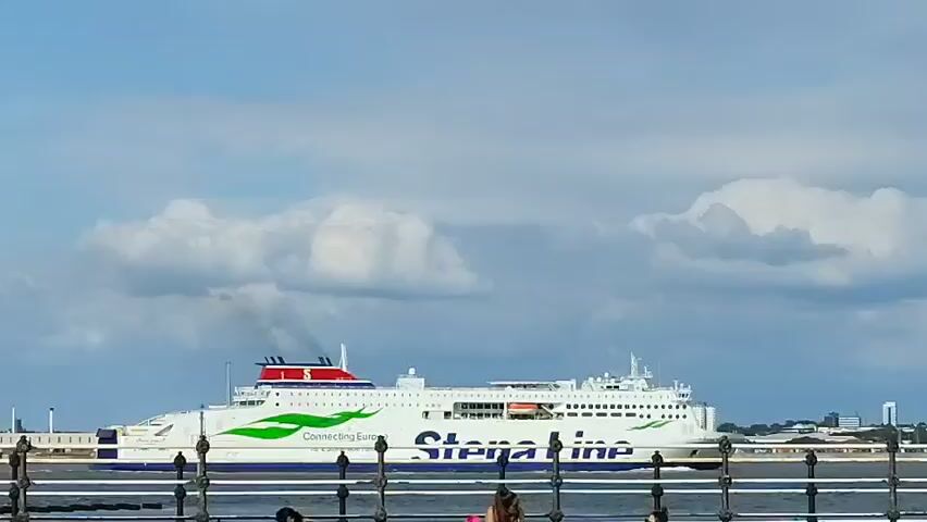 The video shows a large white ferryboat with the Stena Line logo and the words "Connecting Europe" on its side. The ferry is moving from left to right across the frame. Smoke is emitting from a stack on the ferry. The sky is blue with white clouds. The foreground shows a blurry railing and some distant buildings.