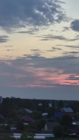 The video shows a village scene at dusk with a wide shot of the sky and a village below. The sky is filled with clouds, some of which are tinted pink from the setting sun. Below, a village with houses and trees is visible. The houses have various colored roofs, including red, blue, and brown. There are also some white structures, possibly greenhouses, in the foreground. The overall atmosphere is peaceful and serene.