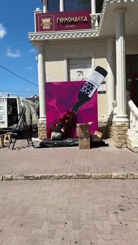 The video shows a person arranging a floral display in front of a building with classical columns and signage in Russian. The floral display is shaped like a large bottle of wine pouring out red and pink flowers and greenery. The person, wearing a black t-shirt, light-colored shorts, and a blue cap, is kneeling and carefully placing flowers. There are cardboard boxes and some loose flowers on the ground around the display. A white van and a ladder are visible in the background, suggesting setup or delivery. The ground is paved with brick-like stones. The sky is blue with some clouds.