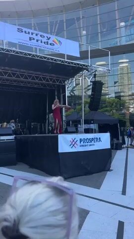 A person in a red sequined dress with a thigh-high slit stands on a stage, holding a microphone and a small object that appears to be a violin bow. They are performing in front of a large, reflective building. A banner above the stage reads "Surrey Pride" with a rainbow graphic. Another banner at the front of the stage displays the "Prospera Credit Union" logo. The person gestures towards the audience, who are partially visible in the foreground, with their hair blonde and styled. The performance appears to be outdoors during the daytime.