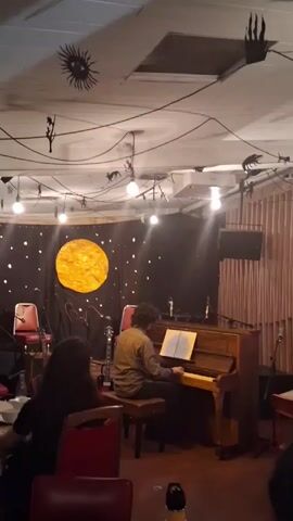 A man is playing a piano in a room. The room has a stage area decorated with a large yellow circle, perhaps representing the moon, against a dark background dotted with white lights.  Two red chairs and a guitar are visible on the stage.  Several black silhouettes of various animals and shapes are suspended from the ceiling by wires along with white lights.  A few people are seated at tables, watching the pianist. A music stand is on the piano.