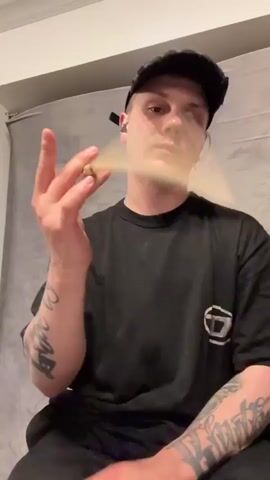 A close-up shot features a man seated and holding a drumstick. He is Caucasian, fair-skinned, wearing a black t-shirt, black cap, and visible tattoos on his arms. He is looking at the camera, his expression neutral. The drumstick is wooden and held in his right hand. The background consists of a grey curtain or backdrop, and a white ceiling is partially visible. The lighting is somewhat dim.