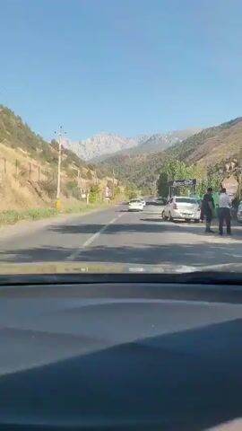 A POV shot from inside a vehicle, driving on a paved road. The road is flanked by a dry, grassy slope on the left and trees and a fence on the right. Mountains with rocky peaks are visible in the distance, under a clear blue sky. Several cars are on the road ahead. A sign with the word "MODERNO" is visible on the right. A person in a red garment is standing near the fence on the right side of the road.