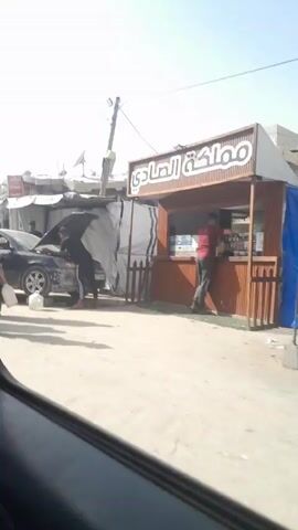 The video shows a scene of war-torn destruction. The view is from inside a car, looking out the passenger window. Initially, people are standing around a black car with its hood open, possibly being worked on. A young boy walks towards the car carrying a white container. As the car moves forward, it reveals a severely damaged building. The building is mostly rubble, with sections collapsed or missing entirely.  A sign with Arabic writing is still visible on the remaining facade of the building. The surrounding ground is dusty and littered with debris. Several individuals are walking around in the vicinity of the damaged building. The sky is clear and blue.