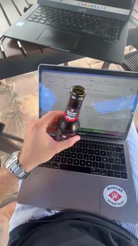 The video begins with a first-person view of someone sitting with a laptop on their lap. A bottle of Estrella Galicia 0.0 alcohol-free beer is held up in front of the screen, obscuring part of the keyboard. A sticker featuring the Detroit Pizza logo is on the lower right corner of the laptop. Another laptop is visible on the table next to it. The camera angle shifts upwards to show a wooden table with two laptops on it, under a covered patio.  The scene then pans out to reveal the patio, complete with a table and chairs, overlooking a swimming pool. Beyond the pool, there's a view of a house and greenery. The setting suggests an outdoor workspace with a leisure area nearby.