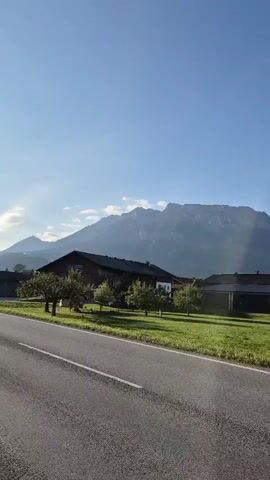 A video pans across a rural landscape under a clear blue sky. In the foreground is a paved road. Beyond the road is a green grassy field with several houses and trees. In the background, a large, hazy mountain range dominates the horizon. The light suggests it is daytime.