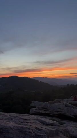 The video shows a group of people sitting on a rocky outcrop, enjoying a sunset view. The sun is setting behind a mountain range, casting a warm orange glow across the sky and clouds. The foreground consists of large, gray rocks, and a forest stretches out beneath the mountains. The overall atmosphere is peaceful and serene.