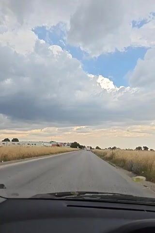 The video shows a view from the interior of a vehicle driving on a paved road. The road is bordered by dry, golden grass on both sides. In the distance, on the left side of the road, there are several buildings with white and red elements, possibly warehouses or industrial structures. Further down the road, a few vehicles are visible. The sky is filled with a mix of dark gray clouds and patches of blue sky with bright white clouds. The overall impression is of a drive through a rural or semi-industrial area under a cloudy sky.