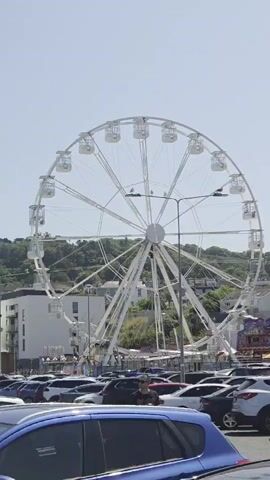 A full shot captures a large white Ferris wheel against a clear blue sky. The Ferris wheel is the central focus of the image, with its numerous cabins visible along its circumference. Below the Ferris wheel, a parking lot is filled with a variety of cars, mostly dark in color, suggesting a public area or attraction. A few people are present, one in the foreground, a man wearing a black t-shirt with a graphic design, and another individual further back, closer to the Ferris wheel. Buildings are visible to the left of the Ferris wheel, and a tree-covered hill forms the backdrop to the right. The overall scene appears to be an outdoor recreational area on a bright, sunny day.