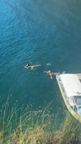 Two women are swimming in a lake. A white boat with a yellow stripe is docked near them. The lake water is a clear turquoise color. In the background, there is a steep hill with trees and small houses on it. The sky is blue.