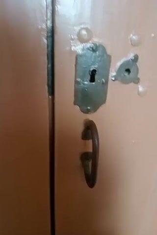 The video begins with a close-up of a weathered door handle being turned and the door opening slightly. The camera then pans to reveal a hallway with a red carpet leading to a set of double doors at the end. To the right of the hallway, a staircase with wooden steps and a railing is visible. The lighting in the hallway appears warm and artificial. The overall environment seems to be indoors, possibly in an older building.