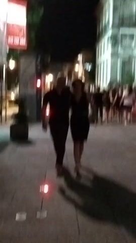 A low-resolution, shaky video captures a street scene at night. A couple walks towards the camera in the foreground, casting long shadows on the paved ground. Behind them, a group of people walks away from the camera. The background shows buildings with lights on. There are streetlights and some red lights visible on the ground. The overall impression is of a lively urban environment at night.