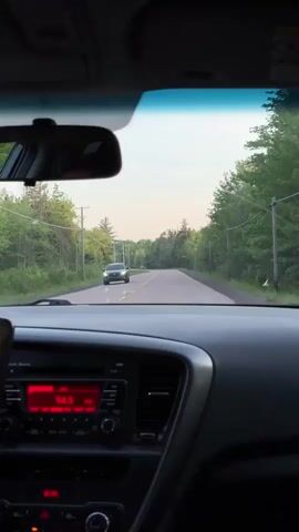 The video is taken from the interior of a car, looking forward through the windshield. The dashboard and radio are visible in the foreground, with the radio displaying "94.5" in red text. A rear-view mirror hangs from the windshield. Outside the car, a two-lane paved road stretches into the distance, bordered by trees and utility poles. The sky is overcast with a pale yellow hue, suggesting either early morning or late afternoon. Another car is visible on the road ahead, traveling in the same direction. The camera appears to be stationary within the car.