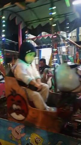 A young child wearing a dark beanie and a light-colored hooded jacket is seated in a small amusement park ride that resembles a train car decorated with cartoon characters. The ride is in motion, indicated by the blurred background and the child's body position. The car has a painting of a red cartoon tomato with a face on its side. The background shows other amusement rides, colorful lights, and trees, suggesting it is nighttime at an amusement park or fairground. The child appears to be looking around and holding onto a steering wheel-like object.