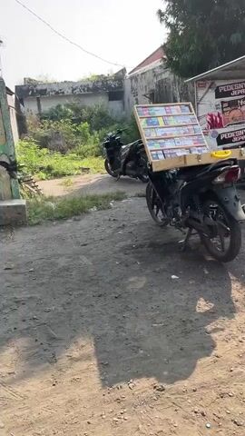 The video shows a street scene with a motorcycle parked on a dirt road. The motorcycle has a wooden display rack attached to the back, filled with what appear to be small items like trading cards or stickers. Another motorcycle is parked behind it. In the background, there are buildings and some overgrown vegetation. The lighting suggests it is daytime.