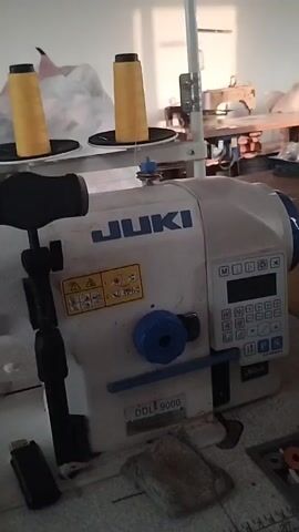 The video shows an indoor scene, likely a workshop or factory, with sewing machines and various supplies. In the foreground, a Juki sewing machine is visible, with two spools of yellow thread on top. To the left of the sewing machine is a black object, possibly a USB drive. Further back, other sewing machines and tables are present. The environment appears to be somewhat cluttered, with blue plastic crates stacked around. The lighting suggests an artificial light source, possibly overhead lighting, casting shadows. The video is shaky, indicating it was recorded by someone holding a handheld device.