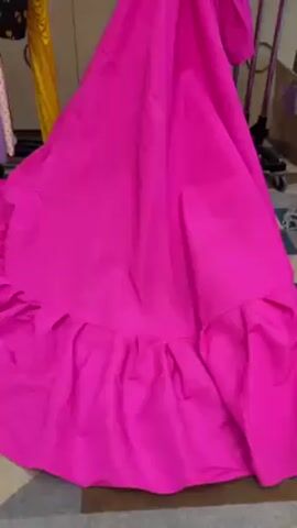 The video shows a brightly lit room with clothing racks and a luggage cart, suggesting it could be a hotel room or a dressing room. A vibrant pink dress with a long train and ruffled hem is displayed prominently on one rack. The room is somewhat cluttered, with cardboard boxes and other items scattered around. In the background, another garment in red is partially visible on a separate rack. The camera pans around the room, focusing primarily on the pink dress and its surroundings. There is no dialogue or human presence in the frame.