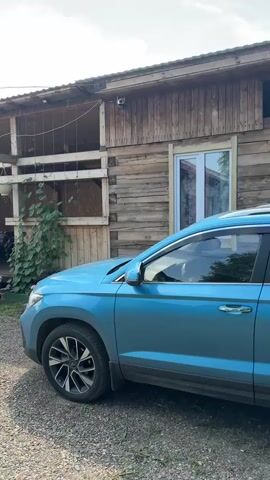 A blue SUV is parked on a gravel driveway in front of a two-story wooden building. The building has a covered porch on the second floor with hay bales stacked on the first floor. There are plants growing up the side of the building. Further down the driveway, a white SUV is parked in front of a smaller building. The area appears to be a rural property with a clear sky and some scattered clouds.