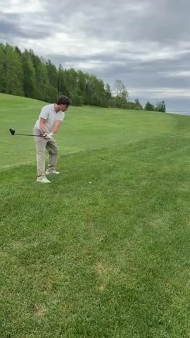 A young man is seen on a golf course. He is wearing a white shirt and khaki pants. The man is swinging a golf club to hit a golf ball on a green grassy hill. There are trees on a hill in the background, and the sky is overcast. After hitting the ball, the man watches it travel down the green.