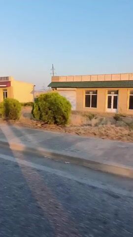 The video shows a moving vehicle's perspective as it travels along a paved road. On the right side of the road, there are two small, low-rise buildings. The first building is a light yellow structure with a red trim and a large glass window. Two bushy green plants are positioned in front of it. The second building is a lighter color with a green roof and a white garage door. Further down the road, a large red and gray metal electricity pylon is visible, with several green trees and bushes surrounding its base. The sky is clear and blue, suggesting it is daytime, and the sun casts shadows across the road. The overall environment appears to be rural or suburban, with dry vegetation and flat terrain in the background.