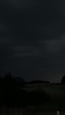 The video shows a dark, overcast sky above a rural landscape. Silhouettes of trees are visible on the left and right sides of the frame. In the center, a gently sloping field or pasture is dimly lit, with a winding road or path visible within it. The overall impression is of a stormy or twilight scene, with very little light. There are no people or overt actions depicted.