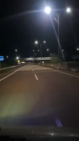 The video shows a car's view of driving on a highway at night. Streetlights illuminate the road. An overpass is visible ahead. The road has lane markings. There is a sign on the left side of the road.