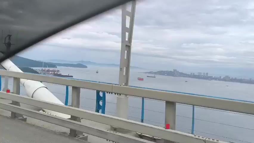 The video begins with a shot of a large white cylindrical object angled upwards against a cloudy sky. A railing with vertical blue elements and horizontal metal bars runs across the bottom of the frame. Beyond the railing, a body of water stretches to the horizon, dotted with several ships. On the left side of the water, a coastline with buildings and a port can be seen. The camera pans slightly to the right, revealing a city skyline in the distance on the right side of the frame. The view is from a high vantage point, likely from a bridge.