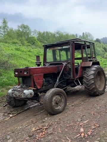 The video shows a red tractor parked on a muddy dirt path. The tractor appears to be old and weathered, with some rust. It has a protective metal cage around the driver's seat. The background includes green vegetation and trees under a cloudy sky. The camera slowly zooms in, focusing on the details of the tractor.